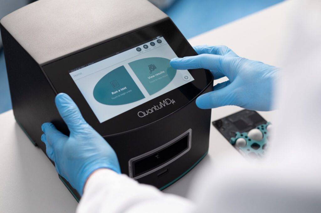 Picture of hands with blue gloves on a Q-POC medical device showing the touch screen with instruction to run a test and see the results